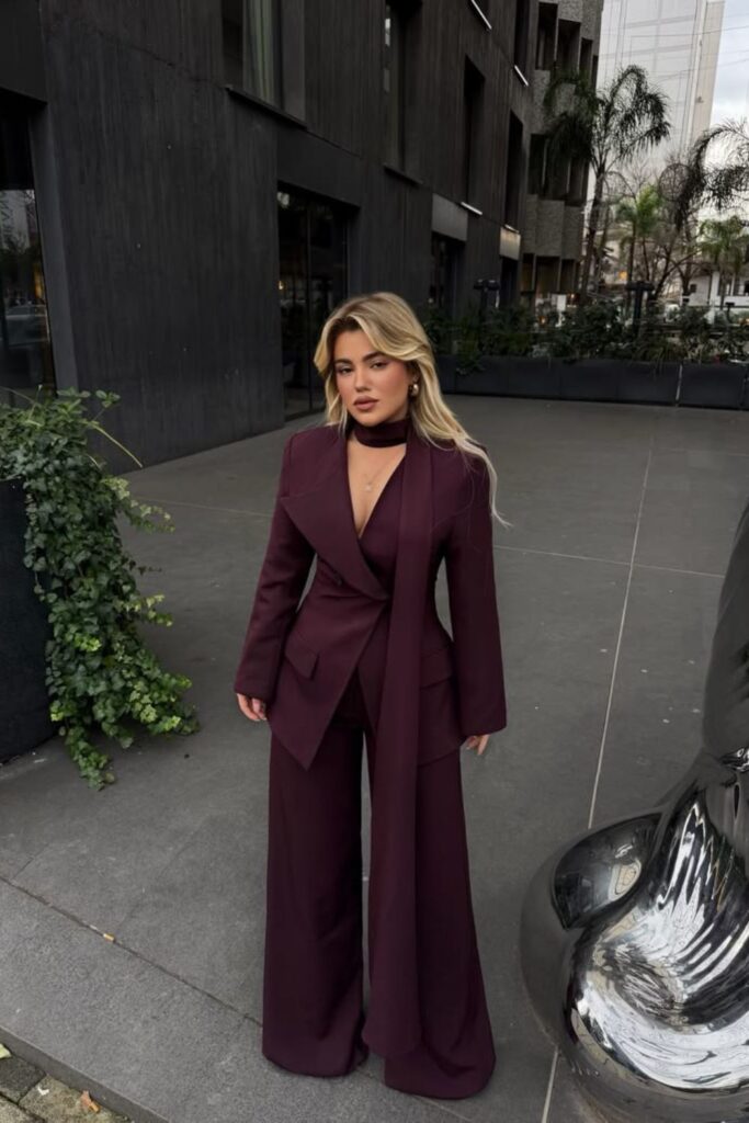 Woman posing outdoors wearing a deep burgundy pantsuit with a matching neck scarf and wide-leg trousers.
