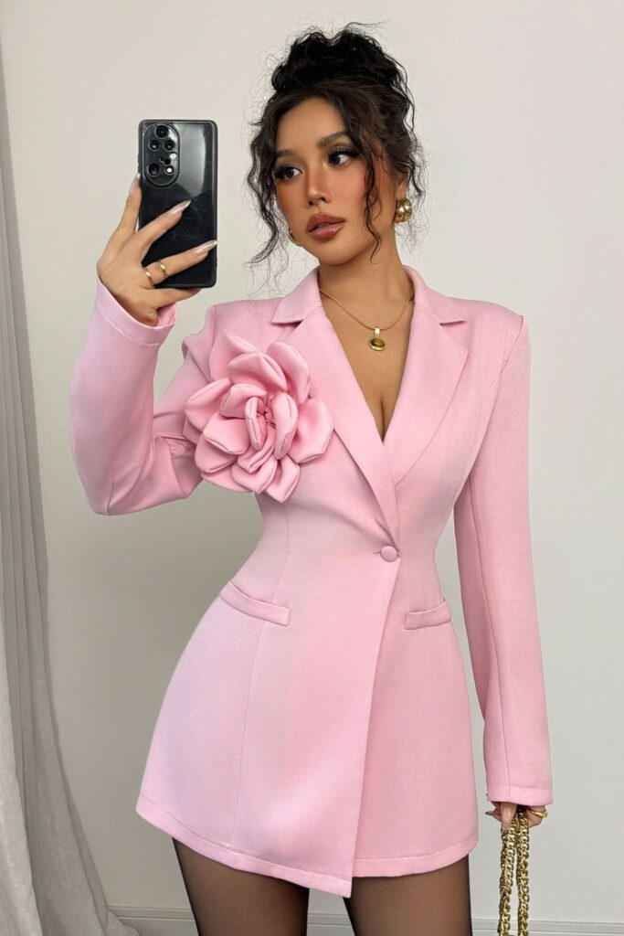 Woman taking mirror selfie wearing a pink long sleeve blazer dress with a large 3D floral decoration.