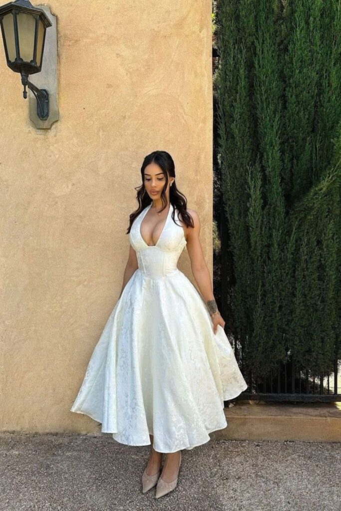 Woman posing outdoors in a white halter neck midi dress with a plunge neckline and a flared A-line skirt.